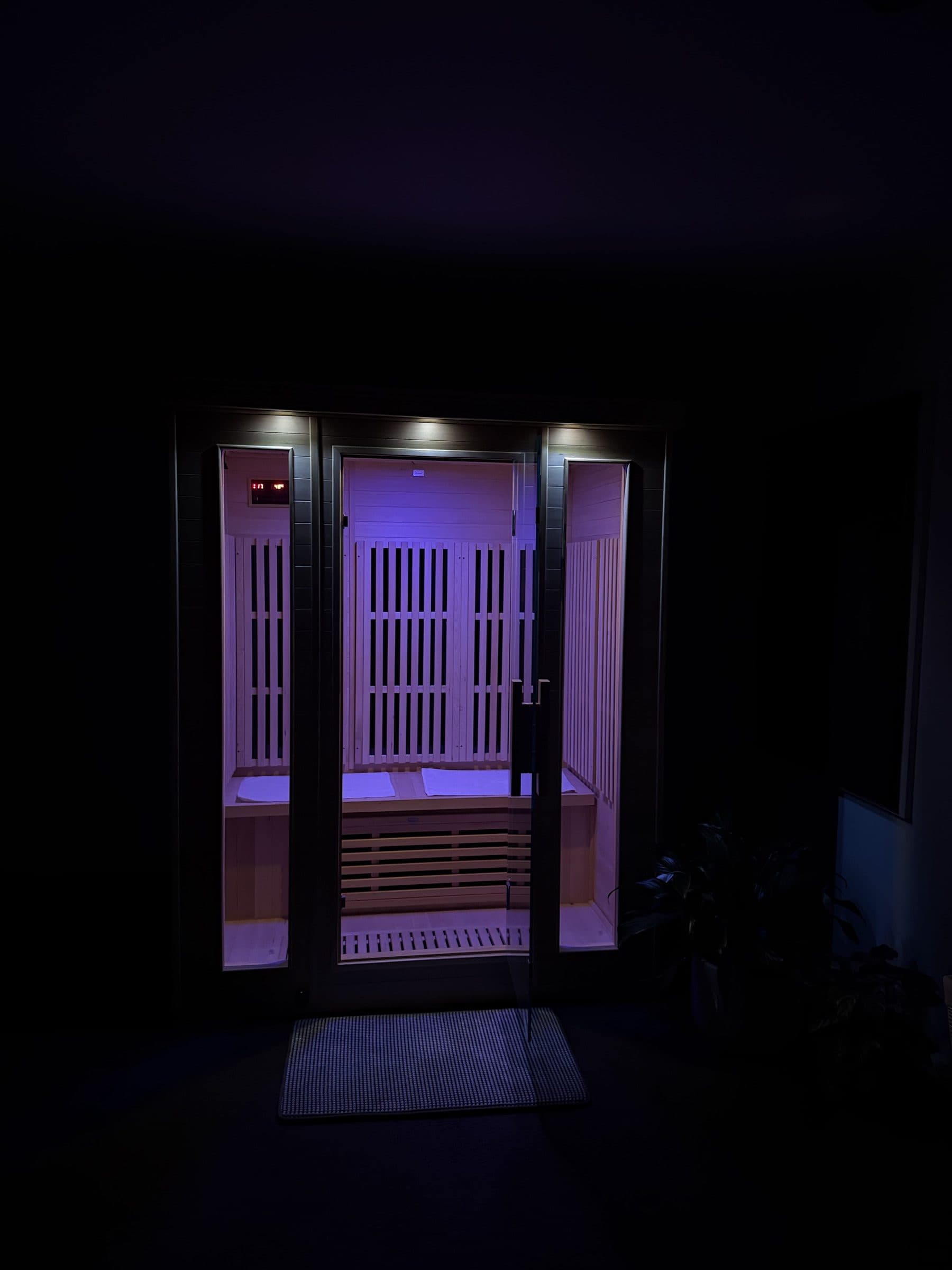 The infrared sauna lit violet by its chromotherapy lamps in a darkened room, its timber interior glowing through the glass doors.